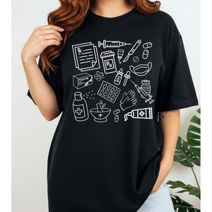 May include: Black t-shirt featuring a white line drawing of medical supplies, including a prescription, syringe, pills, and medicine bottles. The shirt is a comfortable, casual style.
