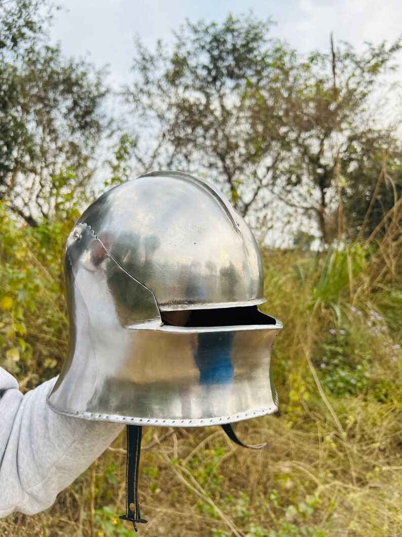 Puede incluir: Un casco medieval pulido, de color plateado, con la parte superior redondeada y una visera. El casco tiene una protecci&oacute;n para el cuello curvada y una correa de cuero con hebilla. El fondo presenta &aacute;rboles y follaje.