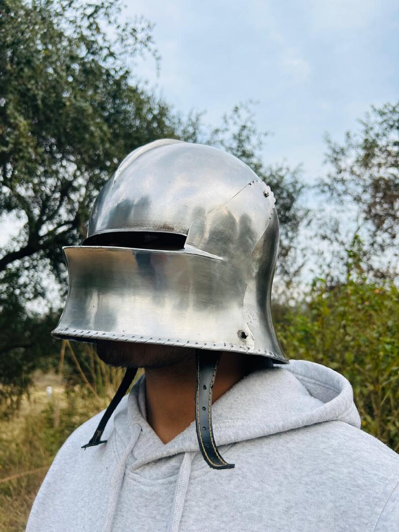 Puede incluir: Un casco medieval pulido de color plateado con visera y correas de barbilla de cuero. El casco est&aacute; dise&ntilde;ado para proteger la cabeza y la cara. El fondo presenta &aacute;rboles y un cielo nublado.