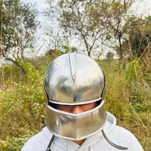 Puede incluir: Un casco de metal plateado pulido con visera y protectores de mejillas. El casco est&aacute; dise&ntilde;ado con una parte superior redondeada y una l&iacute;nea de cejas definida. El fondo presenta follaje verde y &aacute;rboles.