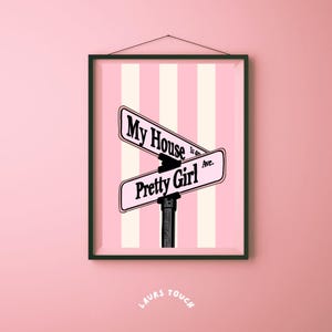 Puede incluir: Arte mural enmarcado con un fondo a rayas rosas y blancas. Dos señales de tráfico negras dicen "My House is on Ave." y "Pretty Girl." La obra de arte está enmarcada en un borde negro y cuelga de una pared rosa.