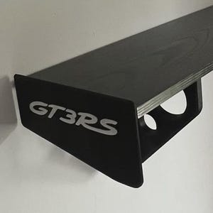 May include: A black wall-mounted shelf with a wood grain texture, featuring side supports with circular cutouts and the white text GT3RS printed on the left side panel. The shelf is designed with a flat horizontal surface and angled brackets.