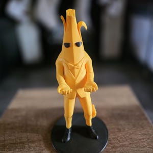 May include: A yellow banana character figurine wearing a yellow suit, black sunglasses, and black shoes. The figurine is on a black circular base. The banana has a bow tie and is posed with its hands open.