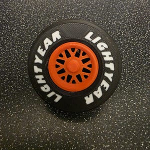 May include: A circular black tyre with a textured surface and a bright orange multi-spoke wheel hub in the centre. The sidewall of the tyre features the white embossed text LIGHTYEAR repeated twice, following the curve of the tyre's outer edge.