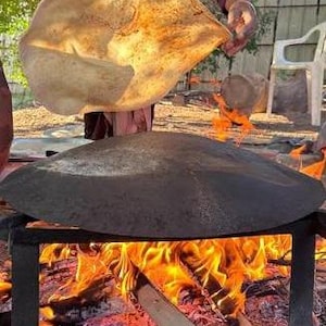May include: A large, round, dark metal griddle sits atop a black metal stand over an open wood fire with bright orange flames. A thin, light-colored, circular flatbread is being lifted from the hot surface of the griddle by hand.