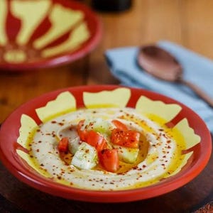 Op de afbeelding: Een close-up van een rode en gele keramische schaal gevuld met hummus, gegarneerd met blokjes tomaten, komkommers en een scheut olijfolie. Een andere schaal met een vergelijkbaar ontwerp staat op de achtergrond.
