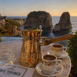 Puede incluir: Una cafetera dorada con un asa de madera y dos tazas de café sobre una mesa de piedra. Las tazas tienen diseños florales. Un cenicero transparente y una servilleta con "CAFE YOUNES" también están sobre la mesa. El fondo muestra una vista panorámica del océano.