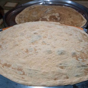 May include: A large, round, flatbread with a light brown color, resting on a dark metal surface. Another similar flatbread is visible in the background, placed on a metal tray. The bread has a slightly textured surface.