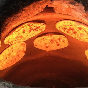May include: A view inside a tandoor oven, showing several pieces of flatbread cooking. The bread is a golden brown colour and has a bubbly texture. The oven interior is illuminated by a bright orange glow.