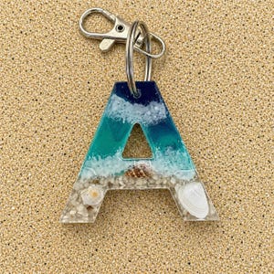 May include: Resin letter "A" keyring with a beach-themed design. The letter features a gradient of blue and teal, with white waves, sand, and seashells. Includes a silver-tone clasp and keyring.