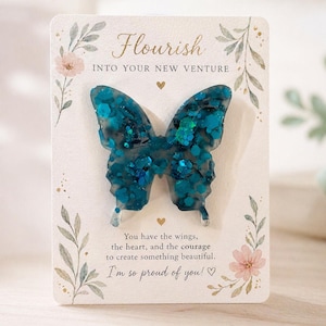 May include: A teal butterfly-shaped resin art piece with glitter accents, presented on a card. The card features the word "Flourish" and the phrase "Into Your New Venture". Decorative floral and leaf elements surround the butterfly.