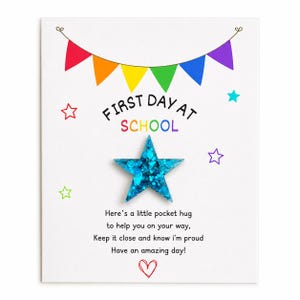 May include: A white card featuring a colorful bunting banner, the text "FIRST DAY AT SCHOOL", and a blue glitter star. Below the star, the text reads: "Here’s a little pocket hug to help you on your way, Keep it close and know I’m proud, Have an amazing day!" with a red heart.
