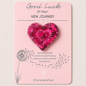 May include: A heart-shaped pin filled with pink and magenta glitter attached to a light pink card. The card features the text Good Luck In Your NEW JOURNEY, a dandelion illustration, and the message The future is bright and full of wonderful things.