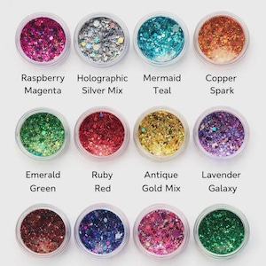May include: A collection of twelve small, clear jars filled with colorful glitter. Each jar is labeled with a color name, including Raspberry Magenta, Mermaid Teal, and Forest Emerald. The glitter colors range from pinks and blues to greens and golds.