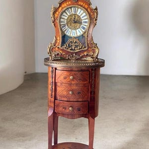 Op de afbeelding: Een hoge houten klok met gedetailleerde goudkleurige metalen ornamenten en een witte wijzerplaat met zwarte Romeinse cijfers. De klok staat op een houten bijzettafel met drie lades, inlegwerk, koperen accenten en een onderste plank.