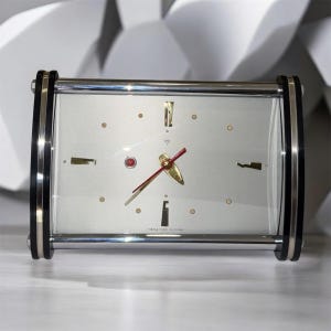 Könnte beinhalten: Eine rechteckige Tischuhr mit silbernem Zifferblatt, goldenen Stundenmarkierungen und Zeigern sowie einem roten Sekundenzeiger. Die Uhr ist in einen Chromrahmen mit schwarzen Seitenteilen gefasst und besitzt eine kleine rote Kontrollleuchte.