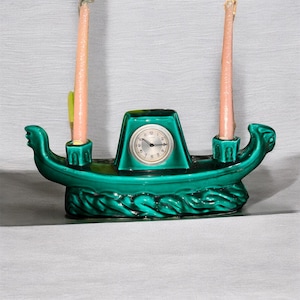 Op de afbeelding: Een teal keramische kaarsenhouder in de vorm van een boot met een klok in het midden. De boot heeft decoratieve golfdetails en twee roze kaarsen. De klok heeft een witte wijzerplaat met zwarte cijfers en wijzers.