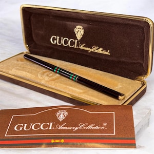 May include: A dark brown Gucci Accessory Collection pen with a gold-tone trim and a green and red striped band. The pen rests inside a brown velvet-lined case with gold-tone edges, featuring the Gucci logo and text printed in gold on the interior.