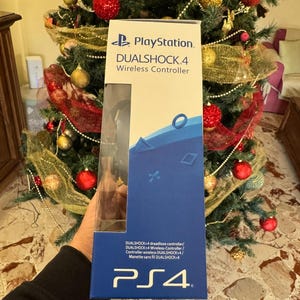 Op de afbeelding: Een blauw en witte verpakking van een PlayStation DualShock 4 Wireless Controller. De verpakking toont het PlayStation-logo, de tekst "DUALSHOCK 4 Wireless Controller" en het PS4-logo. De verpakking wordt voor een versierde kerstboom gehouden.