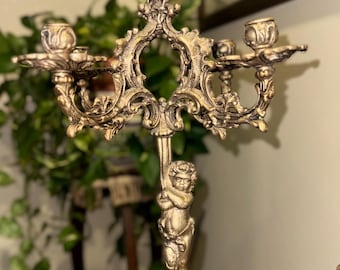 Puede incluir: Un candelabro de metal dorado con una figura central de querubín que sostiene la estructura superior. La pieza tiene tres portavelas con brazos ornamentados y curvos, y una base ancha y decorativa con detalles intrincados de hojas.