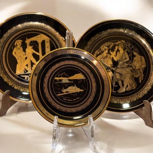 Peut inclure: Trois assiettes décoratives noires avec des motifs dorés de style grec. Les assiettes présentent des scènes de personnages, d'architecture et de paysages. Les assiettes sont exposées sur des présentoirs, mettant en valeur leurs détails et leur thème historique.