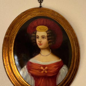 Peut inclure: Un portrait ovale représentant une personne portant un chapeau rouge à larges bords avec un accent jaune, un corsage rouge avec un détail floral et un collier de perles. Le cadre est doré et comporte un ornement fleur de lys au sommet.