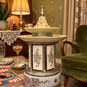 May include: A multi-tiered ceramic carousel music box featuring a cream-colored base and top with hand-painted pink floral motifs. The structure includes silver-toned metal accents, vertical panels with floral designs, and a decorative metal face ornament.