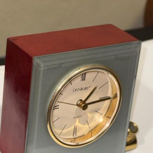 Peut inclure: Une horloge de bureau carrée avec un cadre en bois rouge foncé et une façade en verre dépoli gris. Le cadran circulaire présente une lunette dorée, des chiffres romains noirs et le texte DANBURY et QUARTZ imprimé en noir sur fond crème.
