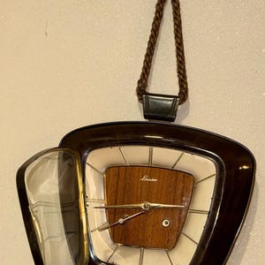 Peut inclure: Une horloge murale avec un cadre brun foncé et un cadran crème doté d'un panneau central en bois avec le texte Linsder. L'horloge possède une porte vitrée articulée sur le côté gauche et est suspendue par un cordon tressé brun avec un ornement.
