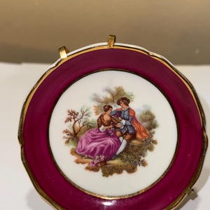 Może przedstawiać: Mały, dekoracyjny talerz porcelanowy z bordową obwódką i złotym wykończeniem. Środek przedstawia malowaną scenę pary w ogrodzie. Talerz jest trzymany przez złoty metalowy stojak.