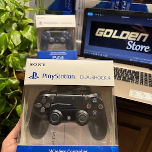 Può includere: Controller wireless DualShock 4 nero per PlayStation, in una scatola di plastica trasparente. La scatola presenta il logo PlayStation e la scritta "Wireless Controller PS4". Un laptop con "GOLDEN Store" è visibile.