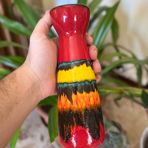 Peut inclure: Un vase en céramique avec un col évasé et un corps cylindrique doté d'un vernis éclatant. La palette de couleurs comprend un haut et une base rouge vif, avec des bandes horizontales jaune, bleu sarcelle et brun foncé en motif goutte.