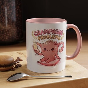 May include: A white ceramic mug with a pink interior and handle, featuring a cartoon illustration of a pink uterus wearing a crown and bow, sitting on a pillow, holding a champagne glass, with the text CRAMPAGNE PROBLEMS printed above.