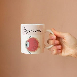 Op de afbeelding: Een witte keramische mok met een grafische illustratie van een menselijk oog in dwarsdoorsnede in rode en roze tinten. Boven de illustratie staat de tekst "Eye-conic" gedrukt in een zwart, handgeschreven lettertype op een lichtroze achtergrond.