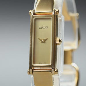 May include: A gold-tone rectangular wristwatch featuring a metallic gold dial with the Gucci logo at the top and the text Swiss Made at the bottom. The watch has a slim, polished gold-tone metal bangle-style strap and a small crown on the right side.