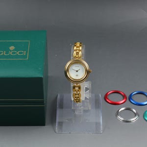 May include: A gold-tone Gucci wristwatch with a white face and a link bracelet, presented with a dark green storage box and five interchangeable colored bezels in red, blue, black, silver, and green, all displayed on a clear stand.