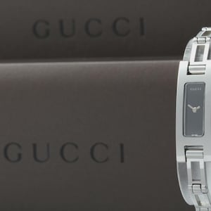 May include: A silver-toned stainless steel wristwatch with a rectangular black dial featuring the Gucci logo. The watch has a polished metal link bracelet and a side crown. The background displays the Gucci brand name in dark brown text.