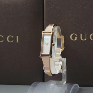 May include: A rose gold-tone Gucci wristwatch with a rectangular case and a white mother-of-pearl dial. The watch features a slim, polished metal bangle-style bracelet and a small crown on the side. The Gucci logo is printed on the dial face.