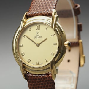 Könnte beinhalten: Eine goldfarbene Fendi-Armbanduhr mit cremefarbenem Zifferblatt und römischen Ziffern. Das Armband ist aus braunem, strukturiertem Leder. Das Zifferblatt zeigt das Fendi-Logo und die Aufschrift "Swiss Made".