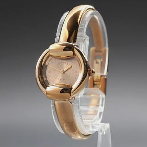 May include: A gold-tone wristwatch featuring a polished round case with a horsebit-inspired design. The dial is a matching gold color with a subtle repeating logo pattern, gold hands, and the text GUCCI and SWISS printed on the face.