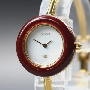 May include: A Gucci wristwatch featuring a round white dial with gold-tone hands, a dark red bezel, and a gold-tone metal bangle-style band. The dial displays the Gucci brand name at the top and the interlocking G logo at the bottom with Swiss Made text.
