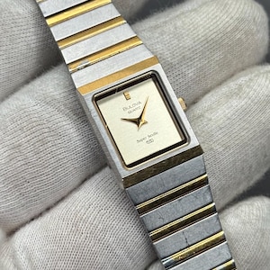 May include: A vintage Bulova Quartz watch with a rectangular gold-toned face and hands. The watch features a silver and gold-toned band. The watch face displays the Bulova logo and the words "Super Seville".
