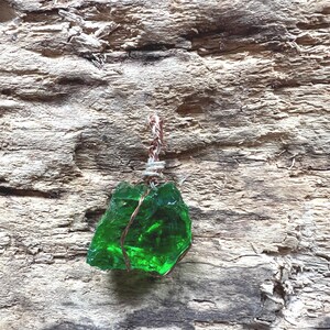 Könnte beinhalten: Ein leuchtend grüner Glasanhänger, unregelmäßig geformt, umwickelt mit Kupferdraht und einer gedrehten silber-braunen Kordel. Der Anhänger ist vor einem strukturierten, verwitterten Holzgrund platziert.