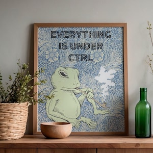 Puede incluir: Lámina artística enmarcada con una rana verde claro fumando en pipa, con el texto "EVERYTHING IS UNDER CTRL" sobre un fondo floral azul. La obra de arte se exhibe sobre una superficie de madera con una cesta tejida, un cuenco de madera y una botella de vidrio verde.