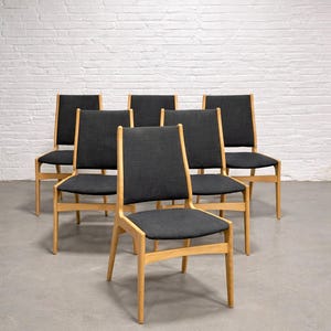 May include: Six wooden dining chairs with black fabric seats and backs. The chairs have a light-colored wood frame and a simple, modern design. The chairs are arranged in a semi-circle, showcasing their clean lines and neutral color palette.