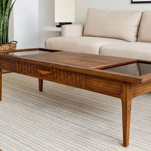 May include: A rectangular wooden coffee table with a glass top and a drawer. The table has a warm brown finish and is supported by four tapered legs. The drawer has a small, triangular handle. The table is approximately 48 inches long.