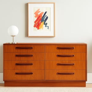 May include: A mid-century modern dresser with eight drawers, crafted from warm-toned wood. The dresser features dark brown, curved drawer pulls and sits on a low base. A framed abstract art piece hangs above the dresser, and a white globe lamp sits on top.