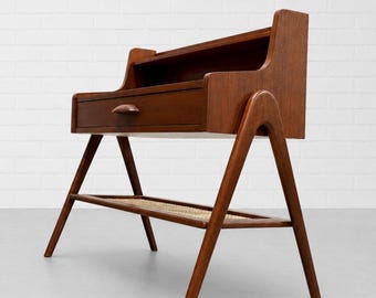 Puede incluir: Una mesa consola de madera marrón con un cajón único con tirador curvo, un estante superior abierto y un estante inferior con superficie de rejilla tejida. La mesa se apoya en cuatro patas inclinadas que forman una estructura en forma de A.
