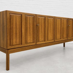 May include: A long, rectangular wooden sideboard featuring a rich brown wood grain finish. The piece has four cabinet doors with vertical panel detailing and small, metallic circular knobs. It is supported by four straight, sturdy wooden legs.