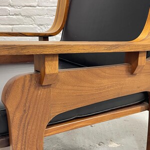 May include: Close-up of a mid-century modern armchair. The chair features a wooden frame with a natural finish and a black leather seat and back. The armrests are curved and the chair has a sturdy, geometric design.